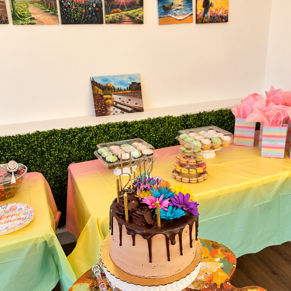 A decorated chocolate drip cake with colorful flower toppers sits on a white pedestal at a decorated event table with pastel tablecloths, macarons, and framed landscape prints on the wall.